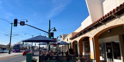 exterior of Que Chevere with customers seated on a patio and a light rail train approaching