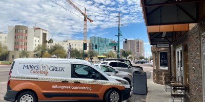 exterior of Mika's Greek with a branded truck used for catering parked outside and high-rise buildings and construction cranes in the backgroun