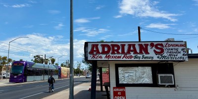 exterior of Adrian's with white signs and red lettering with a light rail train and a bicyclist passing on Main Street