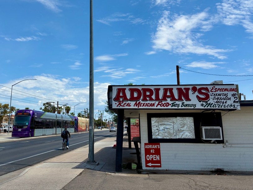 exterior of Adrian's with white signs and red lettering with a light rail train and a bicyclist passing on Main Street