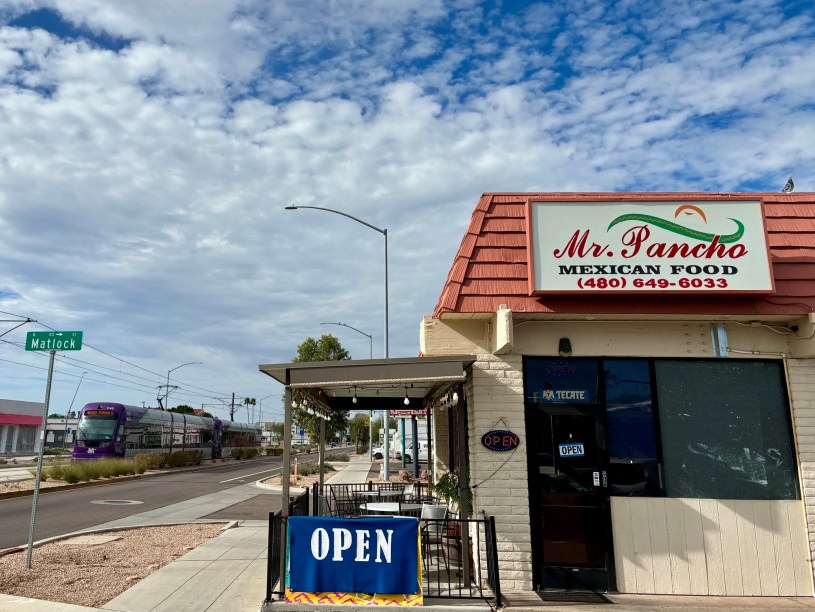 exterior of Mr. Pancho under scattered clouds with a light rail train passing the restaurant on Main Street